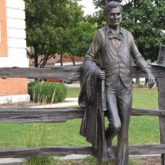 Abraham Lincoln stands next to a split rail fence just outside the courthouse entrance.